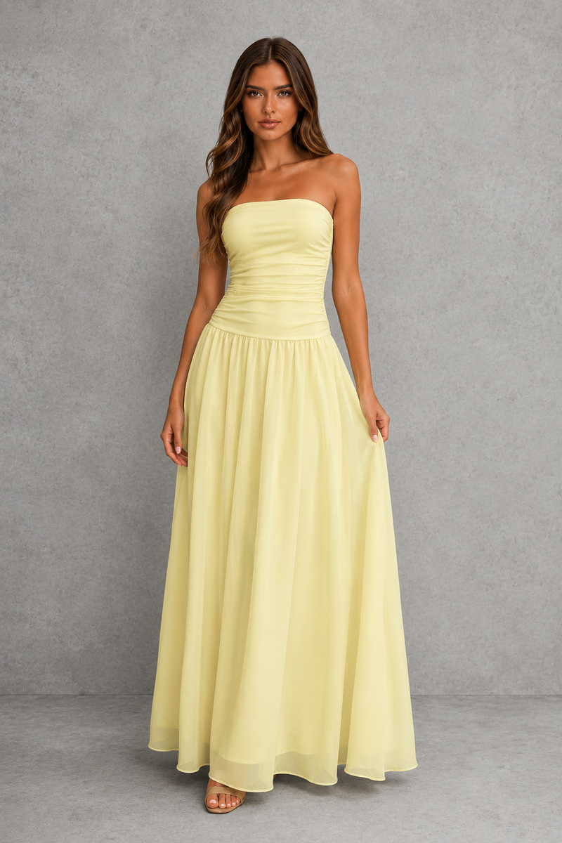 A woman modeling a strapless pale yellow maxi dress with a ruched bodice and flowing skirt, standing against a textured gray studio backdrop.