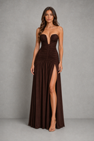 A woman modeling a strapless, floor-length dark brown gown with a plunging neckline, gathered ruching at the waist, and a high front slit, photographed against a neutral gray studio backdrop.