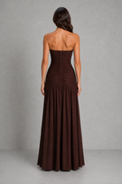 A woman shown from the back wearing a strapless floor-length gown in a very dark brown or near-black color, standing against a plain gray studio backdrop.