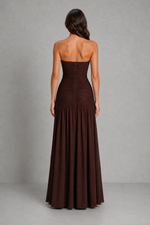 A woman shown from the back wearing a strapless floor-length gown in a very dark brown or near-black color, standing against a plain gray studio backdrop.