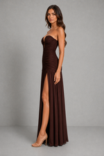 A woman modeling a strapless, dark brown ruched maxi gown with a high thigh slit, standing against a neutral gray studio background.
