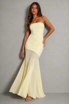 A woman models a strapless pale yellow mermaid gown with a ruched bodice and flowing skirt against a neutral gray studio background.