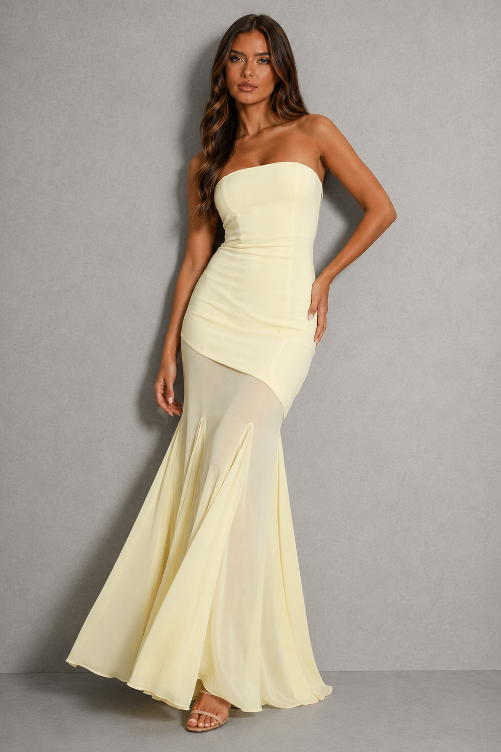 A woman models a strapless pale yellow mermaid gown with a ruched bodice and flowing skirt against a neutral gray studio background.