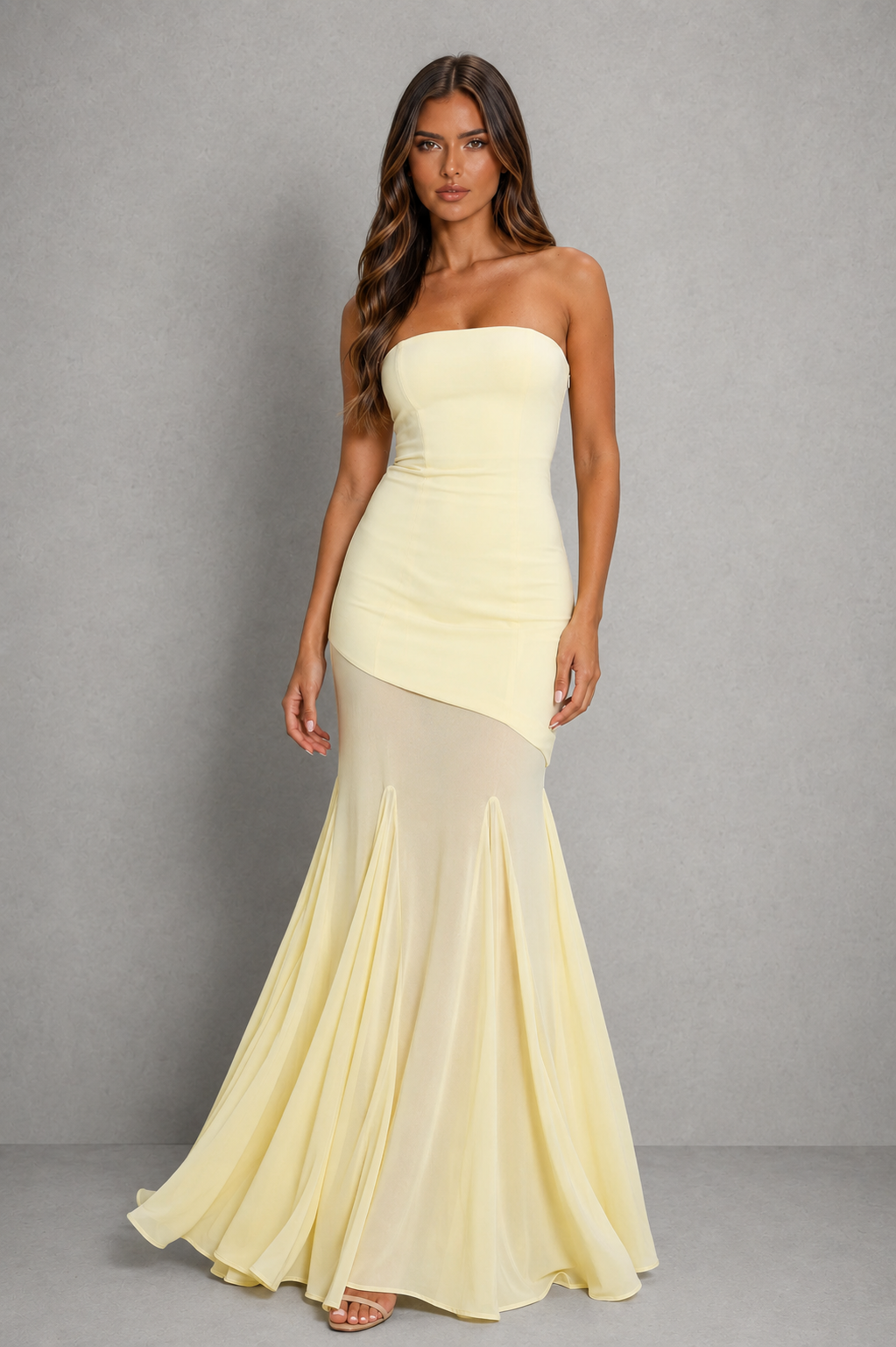 A woman modeling a strapless pale yellow mermaid gown with a structured bodice, asymmetrical draped overlay, and flowing skirt against a plain gray studio background.