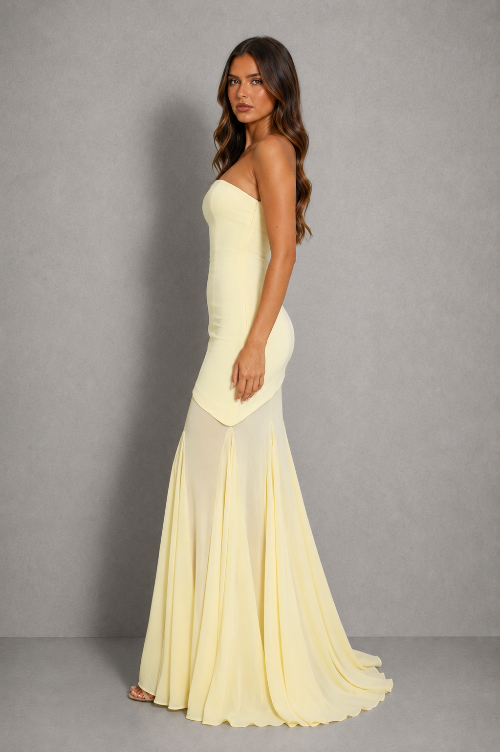 A woman modeling a strapless pale yellow mermaid gown against a neutral gray studio background.