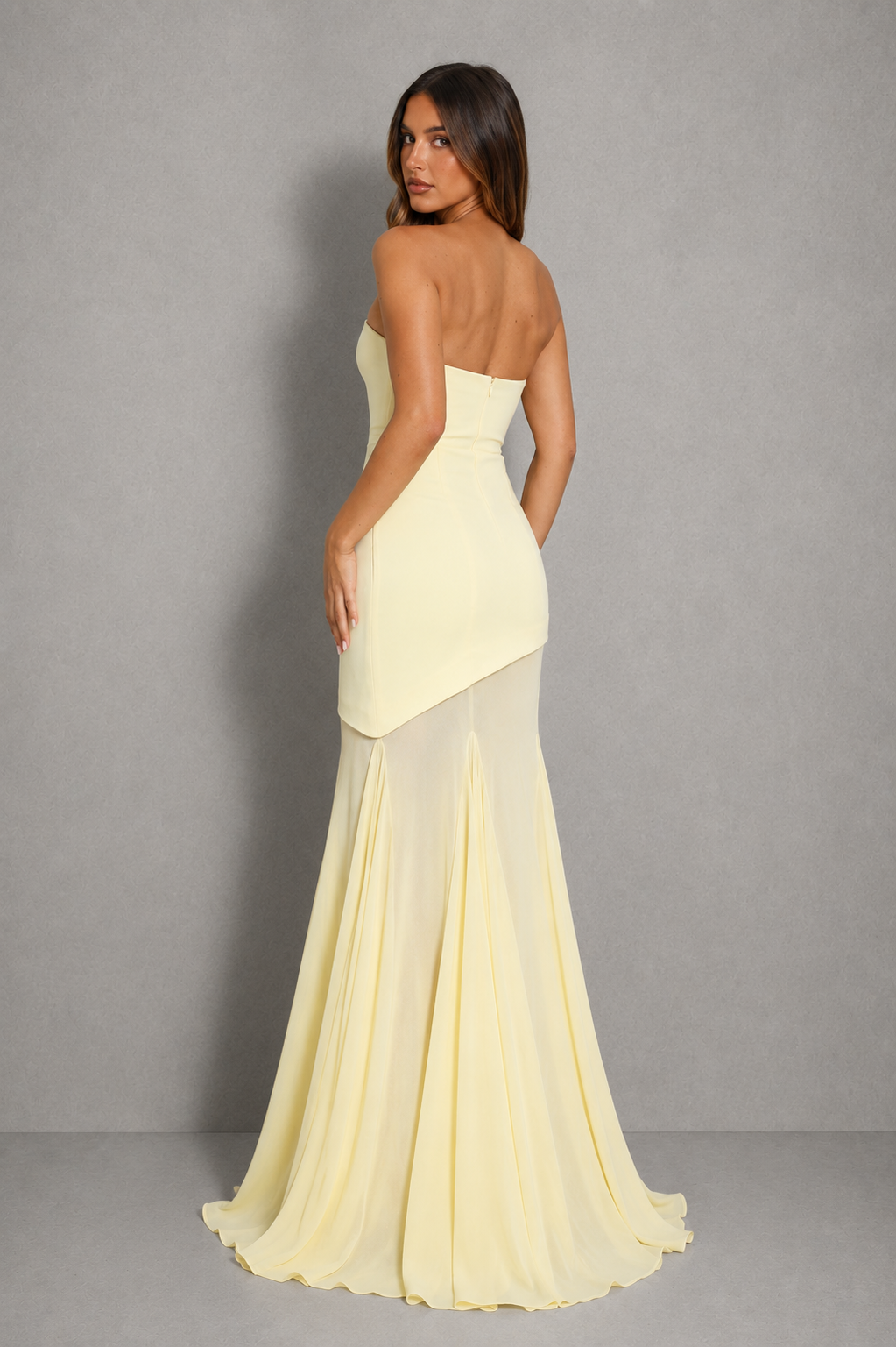 A woman modeling a strapless pale yellow mermaid gown from the back, showing a structured bodice, asymmetrical draped overlay, and flowing chiffon skirt against a gray studio background.