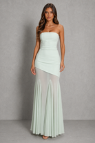 A woman modeling a strapless mint green maxi dress with a structured bodice, asymmetrical draped overlay, and a sheer pleated skirt, set against a textured gray studio backdrop.