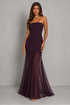 A woman modeling a strapless, floor-length mermaid gown in a deep plum purple color, standing against a textured gray studio backdrop.