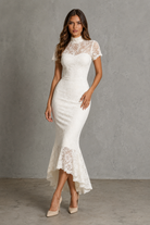 A woman modeling a fitted white lace midi dress with short sleeves, a high mock neck, and a flared lace hem, standing against a textured gray studio wall.