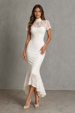 A woman modeling a fitted white lace midi dress with short sleeves, a high mock neck, and a flared lace hem, standing against a textured gray studio wall.