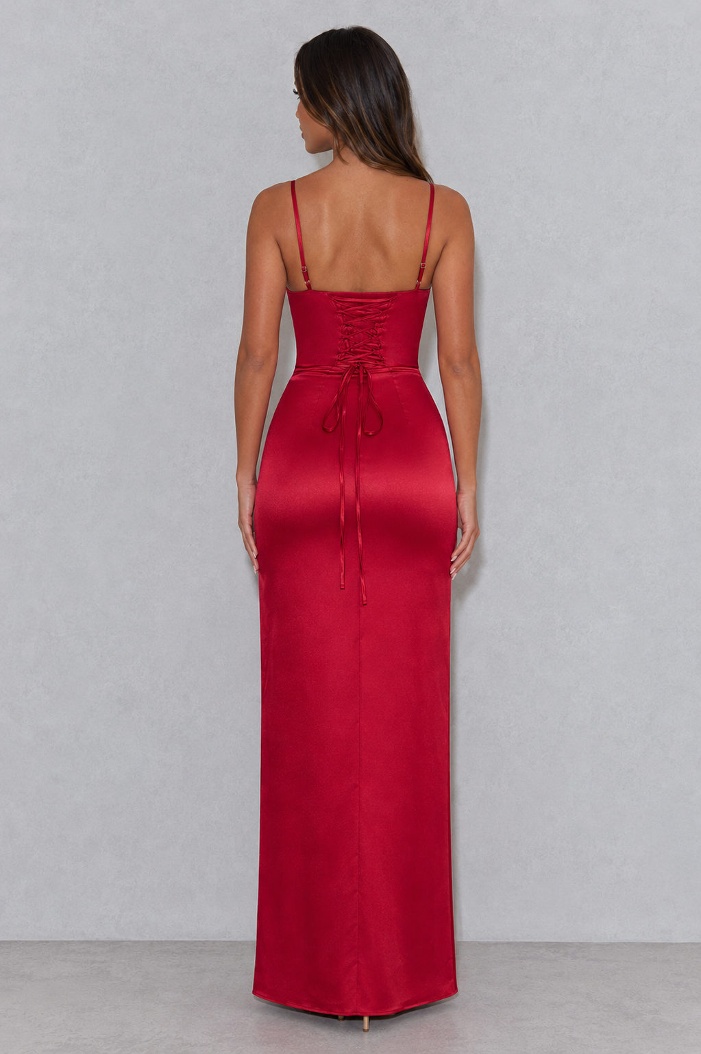 A woman viewed from the back wearing a floor-length red satin dress with thin straps, a lace-up corset-style back, and a tied waist bow, standing against a light gray studio wall.