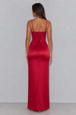 A woman viewed from the back wearing a floor-length red satin dress with thin straps, a lace-up corset-style back, and a tied waist bow, standing against a light gray studio wall.