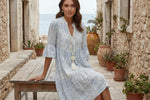 Woman in a light blue patterned dress sitting on a wooden bench with a scenic background.