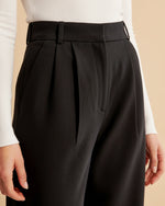 Black pleated pants worn with a white long-sleeve shirt on a beige background