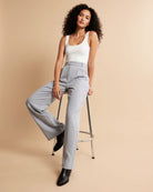 Woman wearing a white tank top and gray high-waisted pants sitting on a stool against a beige background