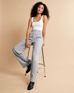 Woman wearing a white tank top and gray high-waisted pants sitting on a stool against a beige background