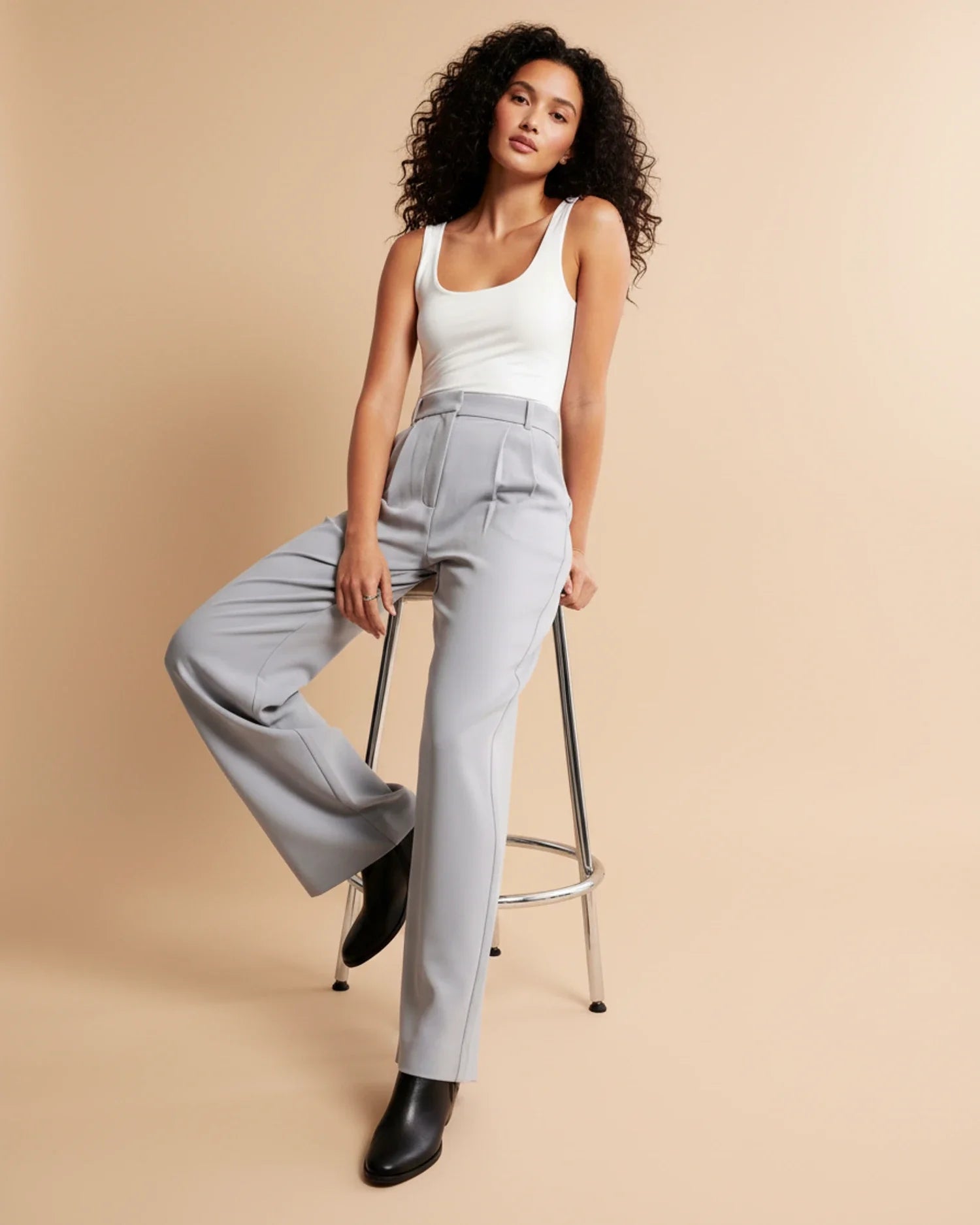Woman wearing a white tank top and gray high-waisted pants sitting on a stool against a beige background
