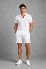 Man wearing a white linen shirt and shorts with white sneakers against a gray background