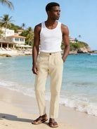 Man in white tank top and beige pants standing on a beach with ocean and palm trees in the background