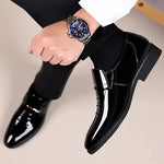 Black dress shoes with shiny finish worn by a person, holding a watch.