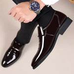 Black shiny dress shoes worn with black pants and a watch on a neutral background