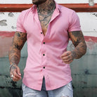 Man wearing a pink short-sleeve shirt with tattoos on arms against a rustic wall.