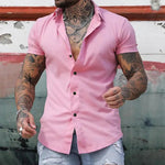 Man wearing a pink short-sleeve shirt with tattoos on arms against a rustic wall.