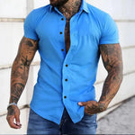 Man wearing a blue short-sleeve shirt with tattoos on arms, standing against a light background.