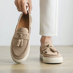 Beige loafers with tassels held by a hand on a light wooden floor.