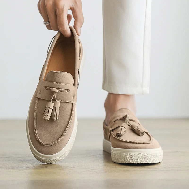 Beige loafers with tassels held by a hand on a light wooden floor.