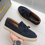 Navy loafers with a beige sole held by a hand on a light gray tiled floor.