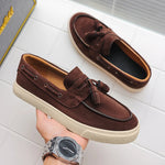 Brown suede loafers with tassels held by a hand wearing a silver watch on a white tiled floor.
