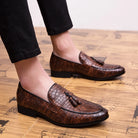 Brown leather loafers with tassels worn by a person on a wooden floor.