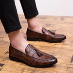Brown leather loafers with tassels worn by a person on a wooden floor.