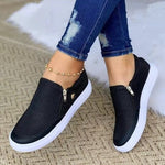 Black slip-on sneakers with white soles worn with blue jeans on a textured surface.