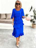 Woman wearing a blue dress in an indoor setting with a couch and plant in the background.