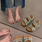 Green sandals with brown soles on a gray floor next to a person wearing blue jeans.