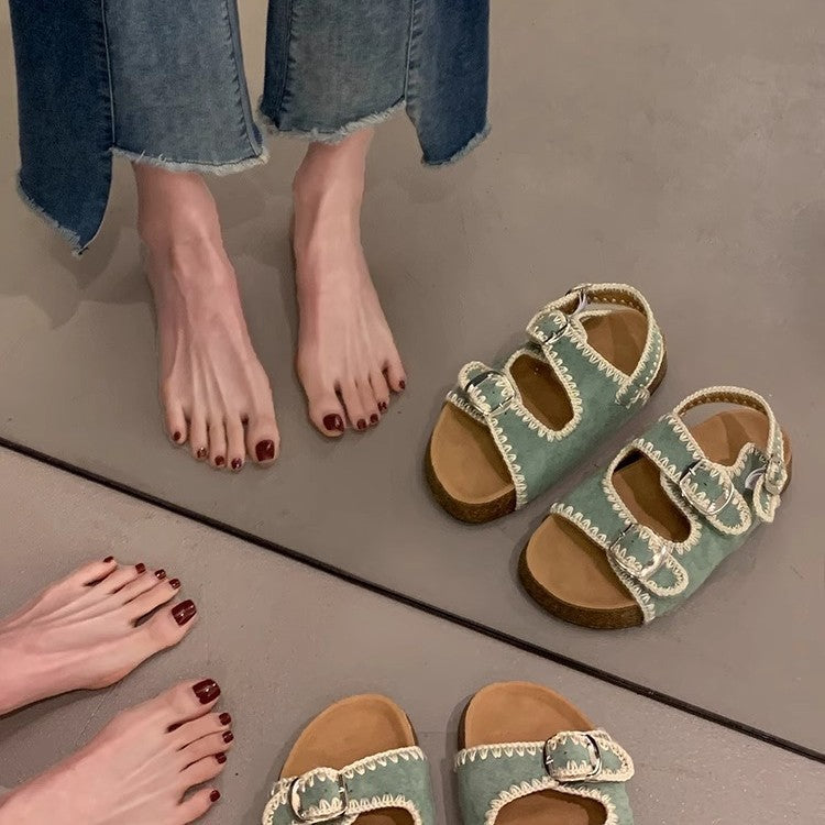 Green sandals with brown soles on a gray floor next to a person wearing blue jeans.