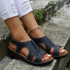 Black sandals worn on feet with white pants and gold anklet, standing on a stone surface.