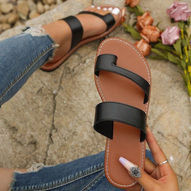 Brown sandal with black straps held by a hand, with jeans and flowers in the background.