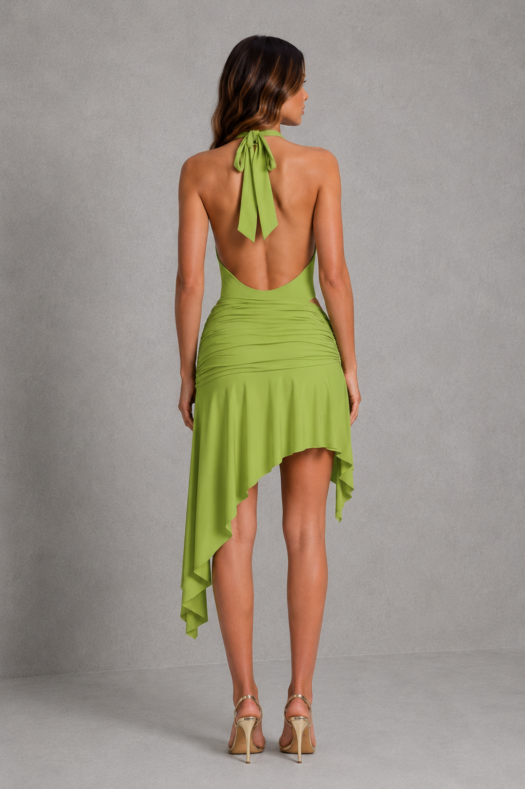 A woman shown from the back wearing a lime green halter-neck dress with an open back and high-low asymmetrical hem, paired with beige high heels, against a gray studio backdrop.