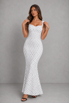 A woman modeling a white sleeveless maxi dress with a black polka dot pattern against a gray studio background.