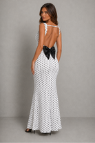 A woman modeling a sleeveless white maxi dress with a black polka dot pattern against a textured gray studio backdrop.