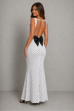 A woman modeling a sleeveless white maxi dress with a black polka dot pattern against a textured gray studio backdrop.