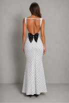 A woman viewed from the back wearing a white sleeveless maxi dress with a black polka dot pattern and a large black bow at the open back, standing against a plain gray studio backdrop.