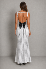 A woman viewed from the back wearing a white sleeveless maxi dress with a black polka dot pattern and a large black bow at the open back, standing against a plain gray studio backdrop.