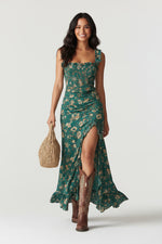 Woman wearing a green floral dress with a high slit and brown cowboy boots, holding a woven bag.