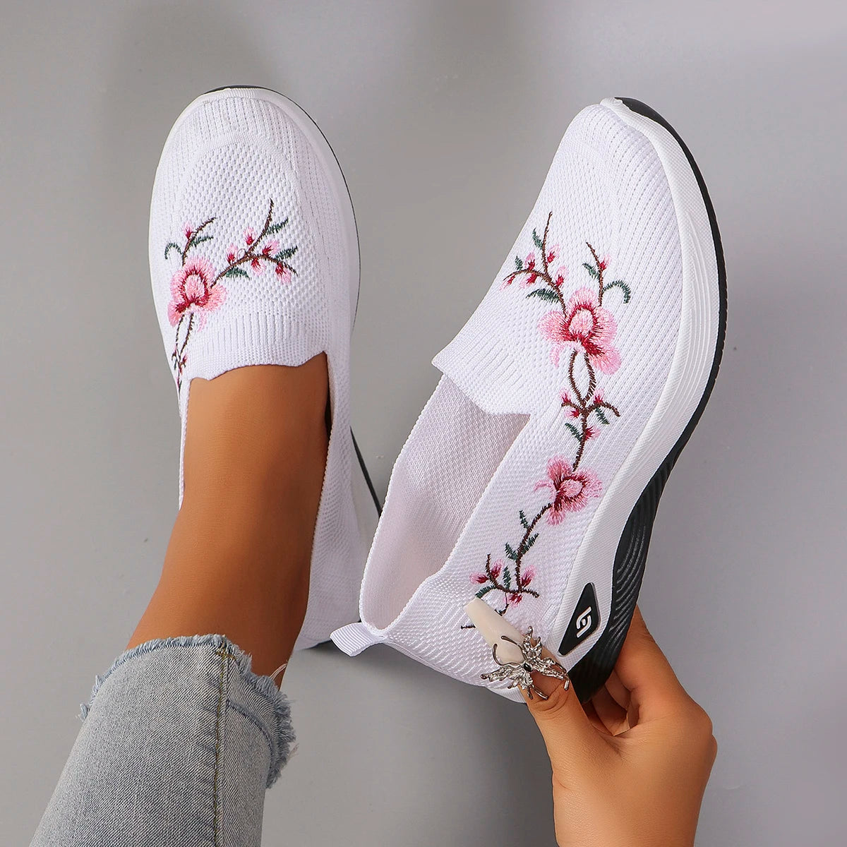 White slip-on shoes with floral embroidery held by a person against a gray background