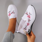 White slip-on shoes with floral embroidery held by a person against a gray background