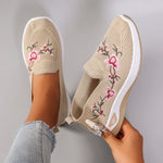 Beige slip-on shoes with floral embroidery held by a hand against a gray background